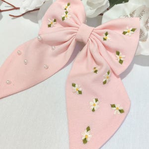 Może przedstawiać: Duża, jasnoróżowa kokarda z haftowanymi białymi i żółtymi kwiatami oraz zielonymi liśćmi. Kokarda ma małe, białe perłowe akcenty i jest wykonana z teksturowanej tkaniny. Ten dodatek do włosów jest idealny, aby dodać odrobinę elegancji.