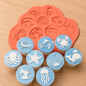 May include: A collection of sea-themed stamps and an orange-red mould. The stamps display white designs of marine life, including a turtle, fish, crab, octopus, starfish, seashell, jellyfish, and whale, on blue fabric circles. The mould has corresponding embossed designs.