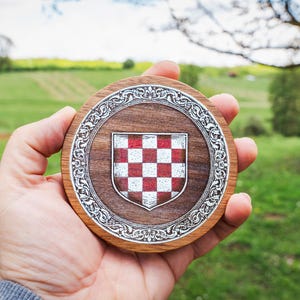 May include: A circular wooden coaster featuring a central shield with a red and white checkered pattern. The shield is surrounded by an intricate silver-toned metal border with ornate scrollwork details set into the dark brown wood grain surface.