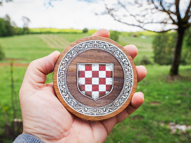 May include: A circular wooden coaster featuring a central shield with a red and white checkered pattern. The shield is surrounded by an intricate silver-toned metal border with ornate scrollwork details set into the dark brown wood grain surface.
