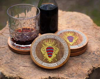 May include: Round wooden coasters featuring a gold and red heraldic crest with a crown and swords. The border has a silver decorative pattern and the text DUBROVNIK at the bottom. One coaster supports a clear glass with a geometric pattern.