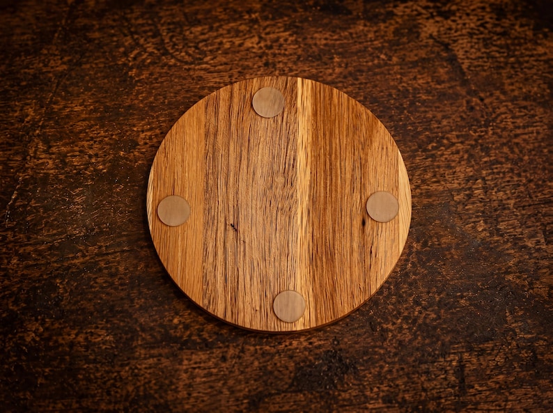May include: A circular wooden coaster featuring a natural light brown wood grain pattern. The underside of the coaster displays four small, circular, light-colored adhesive pads positioned symmetrically near the edge to provide stability.
