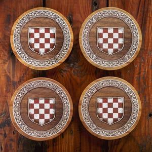 May include: Four circular wooden coasters featuring a central shield emblem with a red and white checkered pattern. Each coaster is surrounded by an intricate, engraved silver-toned border with scrollwork details set against a dark wood grain.