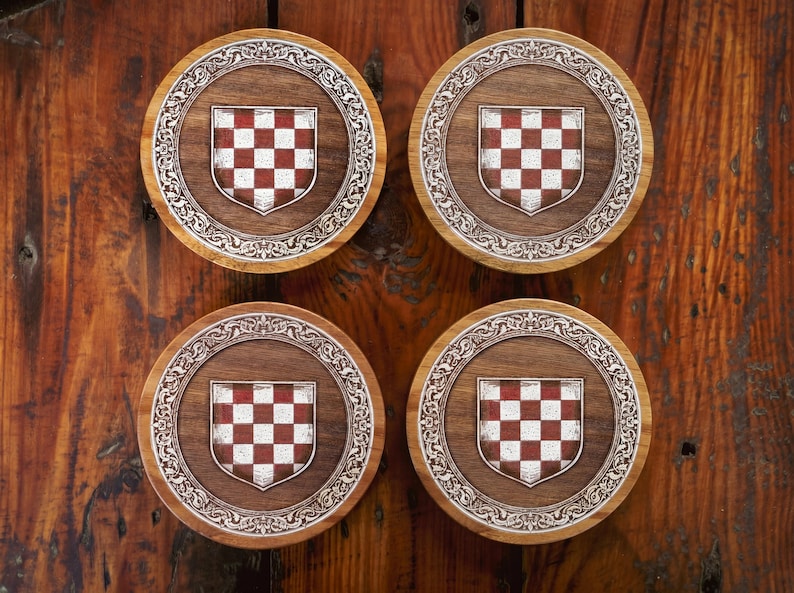 May include: Four circular wooden coasters featuring a central shield emblem with a red and white checkered pattern. Each coaster is surrounded by an intricate, engraved silver-toned border with scrollwork details set against a dark wood grain.