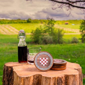 May include: A set of round wooden coasters featuring a central red and white checkered shield emblem surrounded by a silver-toned decorative border, accompanied by a clear glass bottle with a swing-top lid and a crystal-cut drinking glass.