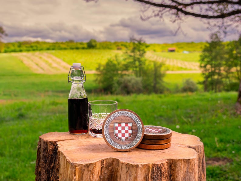May include: A set of round wooden coasters featuring a central red and white checkered shield emblem surrounded by a silver-toned decorative border, accompanied by a clear glass bottle with a swing-top lid and a crystal-cut drinking glass.