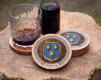 May include: Round wooden coasters featuring a blue shield emblem with three golden lion heads and the text DALMACIJA. The coasters have a dark wood center, a decorative silver-toned border, and a natural wood edge, designed for resting drink glasses.