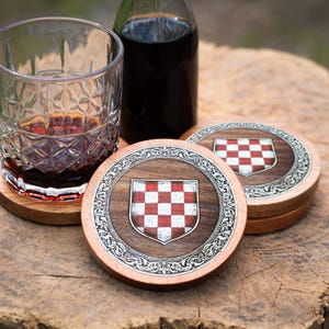 May include: A set of round wooden coasters featuring a central shield emblem with a red and white checkered pattern, surrounded by an intricate silver-toned metal border with embossed scrollwork details, displayed with a glass and a bottle.