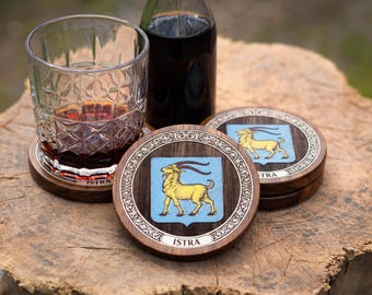 May include: Round wooden coasters featuring a dark brown wood grain border with an intricate silver-toned filigree pattern. The center displays a light blue shield with a yellow goat illustration and the text ISTRA printed at the bottom.