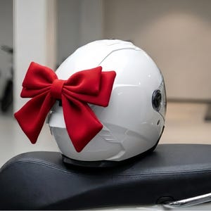 May include: A white motorcycle helmet with a large red bow attached to the back. The helmet is sitting on a black seat, possibly a scooter or motorcycle. The background is blurred, suggesting an indoor setting.