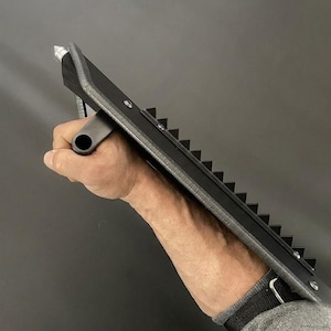 May include: A black and silver tool with a handle and a series of triangular teeth along one edge. The tool has a silver tip and is held in a hand against a dark background.