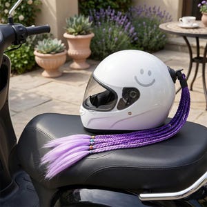 Könnte beinhalten: Ein weißer Motorradhelm mit klarem Visier und einem Smiley-Gesicht. Ein geflochtener Haarteil in Lila mit Perlen ist befestigt. Der Helm liegt auf einem schwarzen Sitz.