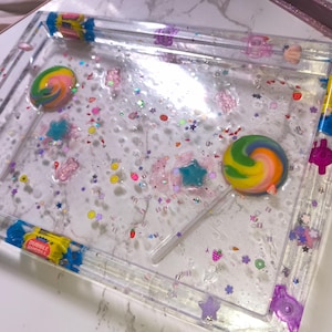 May include: A rectangular clear resin tray featuring embedded colorful swirl lollipops, various small candy-shaped charms, and holographic glitter. The tray edges are decorated with wrapped candy pieces and purple star accents, creating a multi-colored effect.