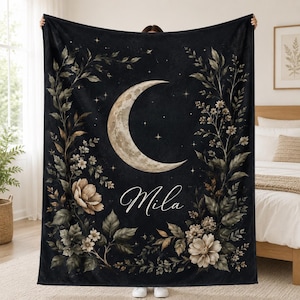 May include: A black rectangular throw blanket featuring a large crescent moon centered at the top, surrounded by botanical illustrations of leaves and flowers. The name Mila is printed in white cursive script below the moon, with small stars.