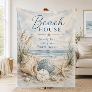 Op de afbeelding: Een rechthoekige plaid met een strandtafereel van schelpen, een zeester en helmgras. De tekst luidt Beach HOUSE, Sandy Feet, Salty Air, Warm Hearts. Het kleurenpalet bestaat uit zachte blauwtinten, zandbeige en witte tinten.