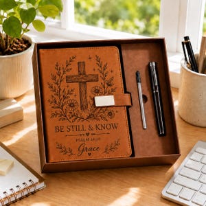 May include: A tan leather-like journal set in a box featuring a cross, floral engravings, and the text BE STILL & KNOW PSALM 46:10 Grace. The set includes a matching pen and a refill cartridge, all secured with a silver-toned metal clasp.