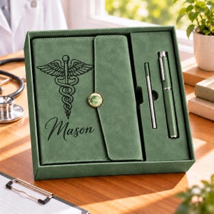 May include: A sage green gift set containing a notebook with a caduceus symbol and the name Mason engraved on the cover, a matching pen, and a thin stylus, all housed in a coordinated green presentation box with a button closure.