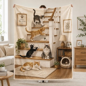 Puede incluir: Una manta de color crema con una ilustración impresa de cinco gatos en una escalera de madera. El texto Home is Where My Cats Are aparece en un cartel enmarcado. Los gatos están representados en varias posiciones, sentados, jugando y tumbados sobre una alfombra.