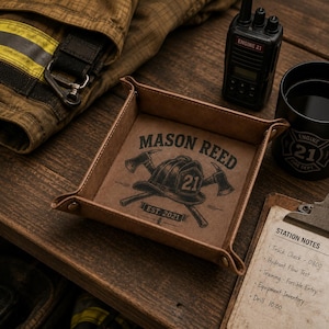 May include: A square tan leather valet tray with snap corners featuring a dark brown graphic of a firefighter helmet, two crossed axes, and the text MASON REED EST. 2021. The tray is constructed with visible stitching along the edges and metal snaps.