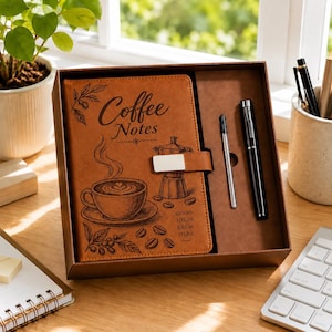 Puede incluir: Un diario de piel sintética color canela en una caja de regalo con un bolígrafo negro y un repuesto. La cubierta presenta una taza de café, una cafetera moka, granos de café y el texto Coffee Notes y Good Ideas Brew Here.