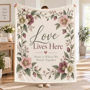 Puede incluir: Una manta de forro polar color crema con un borde floral de flores rosas y blancas con hojas verdes. El texto central dice Love Lives Here con un pequeño corazón y la frase Home is Where We Bloom Together en gris oscuro y granate.