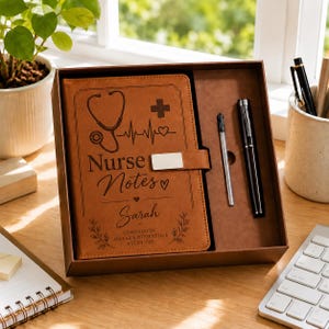 Puede incluir: Un diario de piel sintética color canela con cierre magnético, grabado con un estetoscopio, un corazón, una cruz y el texto Nurse Notes, Sarah, y Compassion Makes a Difference Every Day, presentado en una caja con un bolígrafo negro y recambio.