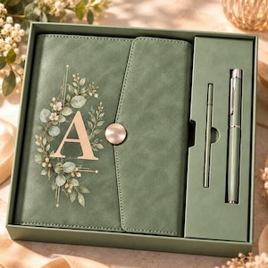 Könnte beinhalten: Ein salbeigrünes Notizbuch-Set bestehend aus einem Journal mit einem goldenen Buchstaben A, umgeben von einem Blumenkranz, und einem passenden Stift. Das Notizbuch hat einen goldenen Knopfverschluss und liegt in einer grünen Box mit Stiftehalter.