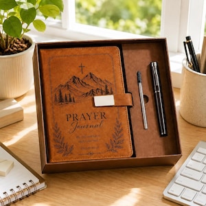 Peut inclure: Un journal de prière en similicuir beige avec un motif de montagne et de croix, portant le texte 'PRAYER Journal', 'Be still, and know that I am God' et 'PSALM 46:10'. Le journal est dans une boîte brune avec un stylo noir et une recharge.