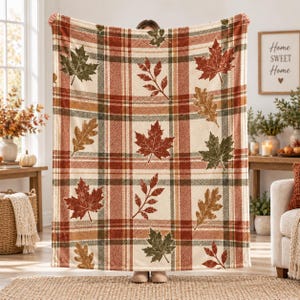 Può includere: Una morbida coperta con uno sfondo color crema e un motivo a quadri marrone, arancione e verde. La coperta è decorata con illustrazioni sparse di varie foglie autunnali, tra cui forme di foglie di acero e di quercia.