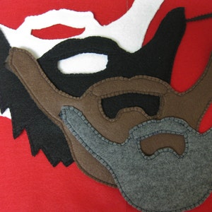 May include: Several felt beard masks in white, black, brown, and grey, displayed on a red backdrop. The masks feature different beard styles and stitched detailing, ideal for fancy dress or theatrical productions.