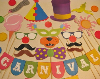 May include: A set of colorful carnival-themed photo booth props. The props include a clown hat, a mask, a mustache, a bow tie, a pair of glasses, a banana, a cotton candy, a sun, and a worm. The props are attached to sticks and are ready to be used for fun photos.