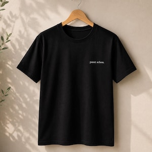 Könnte beinhalten: Ein schwarzes T-Shirt mit kurzen Ärmeln und Rundhalsausschnitt, das an einem Holzkleiderbügel hängt. Auf der Vorderseite befindet sich der kleine weiße Schriftzug 'passt schon.' im oberen rechten Brustbereich in einer schlichten, klaren Schrift.