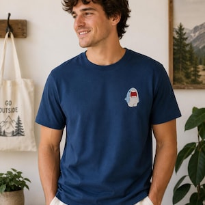 Könnte beinhalten: Ein marineblaues T-Shirt mit kurzen Ärmeln und einer kleinen, mittigen Grafik auf der Brust, die einen hellblauen Cartoon-Hai mit offenem Maul und roten Innendetails zeigt. Das T-Shirt hat einen klassischen Rundhalsausschnitt.