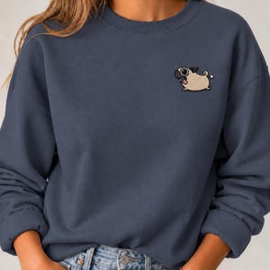 Könnte beinhalten: Ein dunkelblaues Sweatshirt mit Rundhalsausschnitt und einer kleinen Stickerei auf der linken Brust. Das Motiv zeigt einen beigen Mops mit schwarzem Gesicht, großen Augen und geringeltem Schwanz, der rennend mit einer kleinen weißen Wolke dargestellt ist.