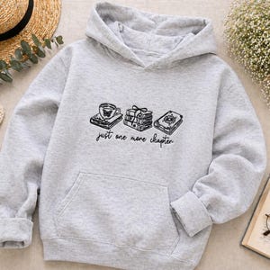 Könnte beinhalten: Ein hellgrauer Kapuzenpullover mit einer schwarzen Strichgrafik, die eine Tasse auf einem Buch, einen Stapel aus drei Büchern und ein einzelnes Buch mit Blumenmotiv zeigt. Unter der Grafik steht der Text "just one more chapter" in schwarzer Schrift.