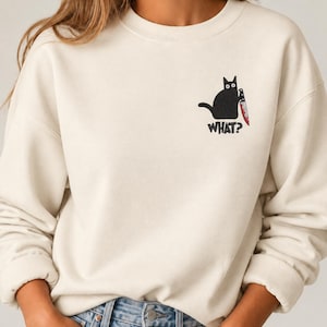 Könnte beinhalten: Ein cremefarbenes Sweatshirt mit Rundhalsausschnitt und einer Stickerei auf der linken Brust. Das Motiv zeigt eine schwarze Katze, die ein Messer mit roter Klinge hält, ergänzt durch den Schriftzug "WHAT?" in schwarzen Buchstaben darunter.
