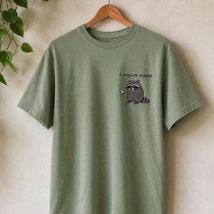 Könnte beinhalten: Ein salbeigrünes Kurzarm-T-Shirt mit einer Stickerei auf der linken Brust, die einen Waschbären mit einem kleinen Messer zeigt. Über dem Waschbären steht der Text i require snacks in einer schwarzen Schriftart im Schreibmaschinen-Stil.