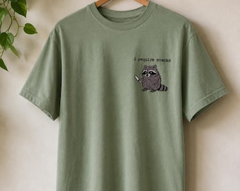 May include: A sage green short-sleeve t-shirt featuring an embroidered graphic on the left chest of a raccoon holding a small knife. Above the raccoon, the text reads i require snacks in a black typewriter-style font. The shirt has a crew neck.