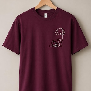 May include: A maroon short-sleeved t-shirt featuring a white line art graphic on the left chest. The design depicts a stylised dog silhouette formed by a single continuous line, incorporating a small heart shape near the dog's nose and chest.
