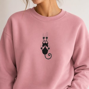 Könnte beinhalten: Ein altrosa Sweatshirt mit Rundhalsausschnitt und einer schwarzen Stickerei, die eine Katze zeigt, die sich mit ihren Krallen festhält. Die Katze hat große Augen und einen geringelten Schwanz, mit vertikalen Linien über den Pfoten.