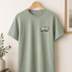 Könnte beinhalten: Ein salbeigrünes Kurzarm-T-Shirt mit einer kleinen Grafik auf der linken Brustseite. Die Grafik zeigt eine liegende Glückskatze mit dem Text "NOT TODAY", der in schwarzen Großbuchstaben direkt unter der Katzenillustration gedruckt ist.