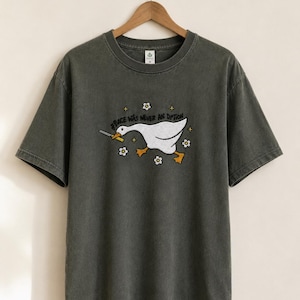 Könnte beinhalten: Dunkelgraues T-Shirt mit einem humorvollen Motiv einer weißen Gans, die ein Messer hält, umgeben von kleinen Blumen. Der Text "Peace Was Never An Option" steht über der Gans. Das T-Shirt hängt an einem Holzbügel.