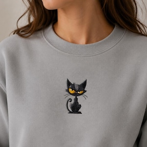 Könnte beinhalten: Ein hellgraues Sweatshirt mit Rundhalsausschnitt und einer kleinen, mittigen Stickerei einer schwarzen Katze mit großen, leuchtend gelben Augen und einem mürrischen Ausdruck. Die Katze hat einen dünnen, geschwungenen Schwanz und spitze Ohren.