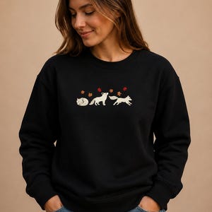 Könnte beinhalten: Ein schwarzer Rundhals-Pullover mit einer Stickerei von drei weißen Füchsen in verschiedenen Posen, begleitet von kleinen roten und orangefarbenen Herbstblättern, die darüber verstreut sind. Der Pullover hat lange Ärmel sowie gerippte Bündchen.