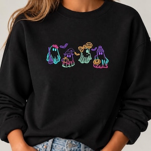 Könnte beinhalten: Ein schwarzes Sweatshirt mit Rundhalsausschnitt und einer bunten Neon-Grafik von vier Geistern. Die Geister sind mit verschiedenen Accessoires wie einem Besen, einem Hexenhut, einer Schleife und einer Mütze sowie kleinen Fledermäusen dargestellt.