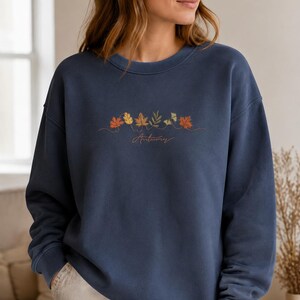 Könnte beinhalten: Ein marineblaues Sweatshirt mit Rundhalsausschnitt und einer Stickerei aus verschiedenen Herbstblättern in Orange-, Gelb- und Grüntönen, wobei der kursive Schriftzug "Autumn" in dünnem braunem Garn unter dem Motiv platziert ist.