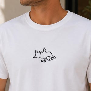 Könnte beinhalten: Ein weißes T-Shirt mit kurzen Ärmeln und einer kleinen schwarzen Stickerei auf der Brust. Das Motiv zeigt eine Katze, die auf dem Rücken liegt und eine Pfote hebt, begleitet von dem in schwarzem Garn gestickten Text NÖ unter der Katze.