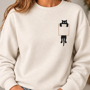 Könnte beinhalten: Ein cremefarbenes Sweatshirt mit Rundhalsausschnitt und einer Stickerei auf der linken Brust. Die Stickerei zeigt eine schwarze Katze mit weißen Augen, die oben aus einer rechteckigen Taschenkontur herausragt, wobei der Schwanz nach unten hängt.