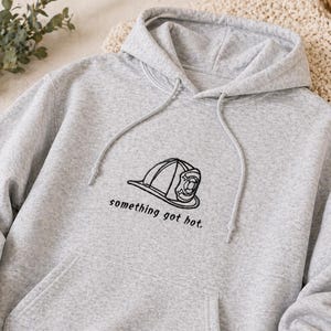 Könnte beinhalten: Ein hellgrauer Kapuzenpullover mit Kordelzug und Vordertasche. Der Pullover hat ein schwarzes gesticktes Design eines Feuerwehrhelms und den Schriftzug "something got hot."