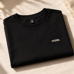Könnte beinhalten: Ein schwarzes kurzärmeliges T-Shirt mit Rundhalsausschnitt. Auf der rechten Brustseite ist das Wort "müde." in hellem Beige aufgedruckt. Am Halsausschnitt befindet sich ein kleines schwarzes Etikett mit einem weißen Grafiksymbol und dem Buchstaben M.